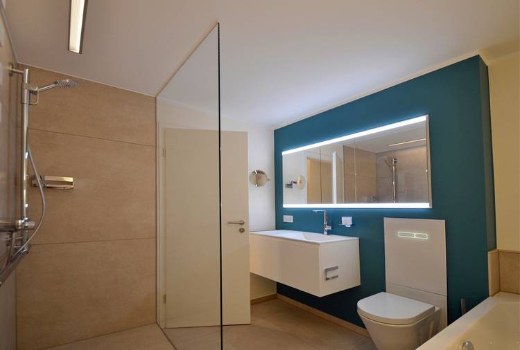 Modernes Badezimmer mit Glasdusche, wandmontiertem Waschbecken, hinterleuchtetem Spiegel und Toilette auf hell gefliestem Boden.