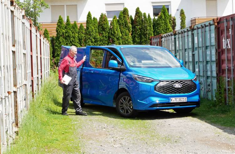 Alt-Text: „Fachhandwerker im roten Hemd begutachtet Baupläne neben einem blauen Servicefahrzeug zwischen Frachtcontainern auf einer Baustelle für Gebäudetechnikprojekte.