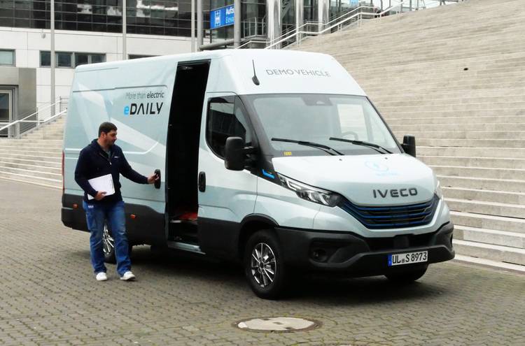 ALT-Text: „Ein Fachhandwerker begutachtet einen geparkten IVECO eDAILY Elektro-Van vor einem modernen Gebäude, um die Integration von nachhaltigen Mobilitätslösungen in zukunftsorientierte Gebäudekonzepte zu evaluieren.