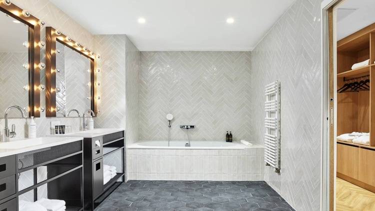 Alt-Text: „Modernes Badezimmerkonzept mit doppeltem Waschbecken und Waschtisch, freistehender Badewanne und begehbarem Kleiderschrank. Die Gestaltung mit grauen Wandfliesen und hexagonalen Bodenfliesen bietet ein Beispiel für energieeffiziente Beleuchtung und nachhaltige Materialien im Sanitärbereich.