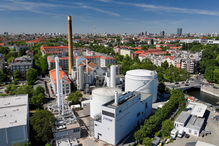 ALT-Text: Luftaufnahme einer modernen Industrieanlage mit Rauchgasreinigungsanlagen, umgeben von städtischer Bebauung und Grünflächen, illustriert das Thema nachhaltige Energieversorgung in der Gebäudetechnik unter klarem blauen Himmel.