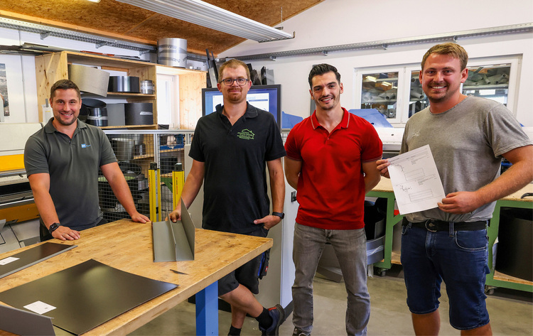 ALT-Text: „Vier Fachleute der Gebäudetechnik diskutieren in einer Werkstatt über Pläne für ein energieeffizientes Heizungs- und Kühlsystem, umgeben von Metallplatten und Werkzeugen.