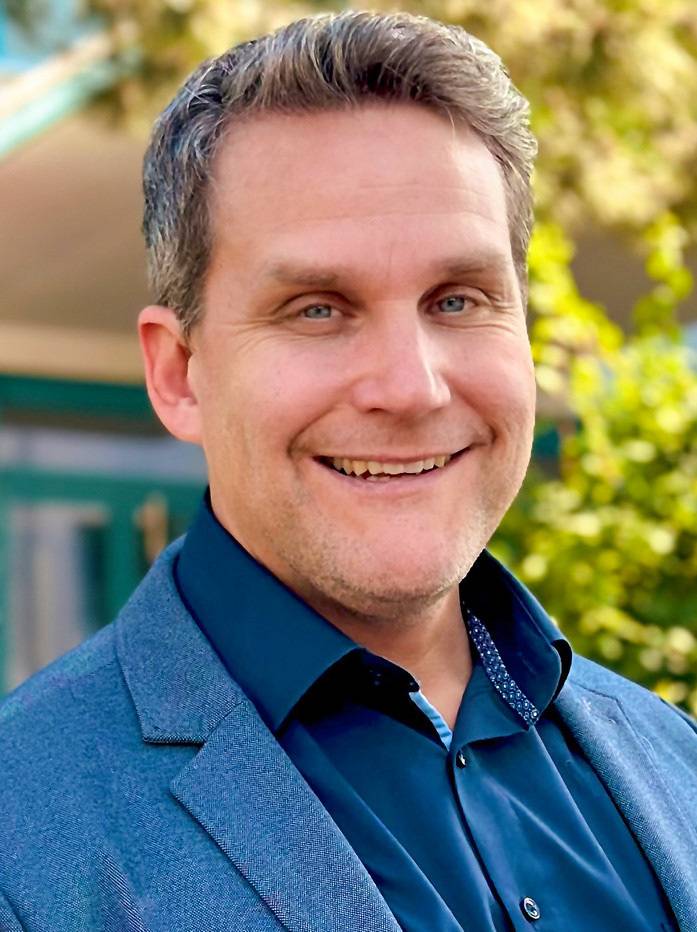 ALT-Text: „Fachkundiger Gebäudetechnik-Experte in blauer Arbeitskleidung, lächelt im Freien vor einer Kulisse voller Pflanzen, symbolisiert Engagement für Nachhaltigkeit und Energieeffizienz in der modernen Gebäudetechnik.