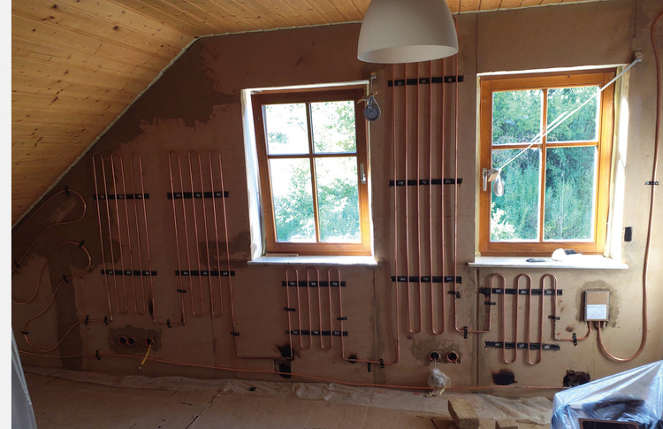 Alt-Text: „Verlegte Kupferrohre eines Fußbodenheizungssystems an einer renovierten Innenwand mit Fenstern, exemplarisch für nachhaltige Heiztechnik in modernen Bauprojekten.