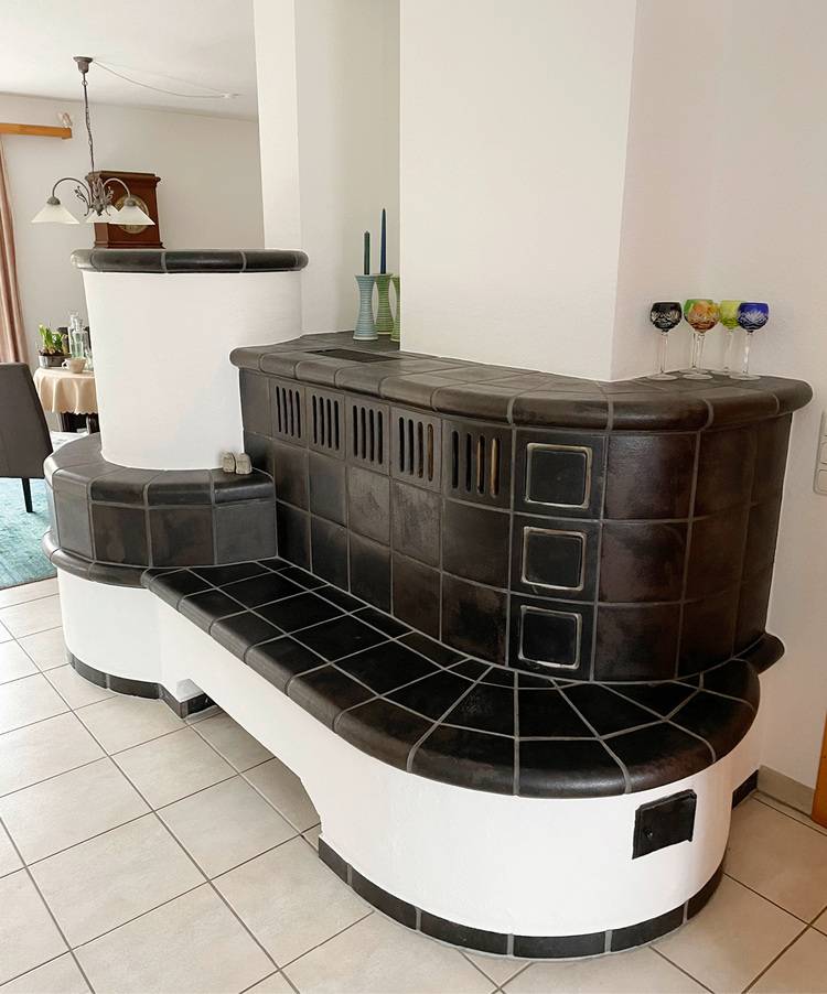 ALT-Text: „Effizienter, schwarz gekachelter Heizofen mit integrierter Sitzbank in einem hellen Raum, ideal für moderne Gebäudetechnikkonzepte und Energieeffizienzlösungen.