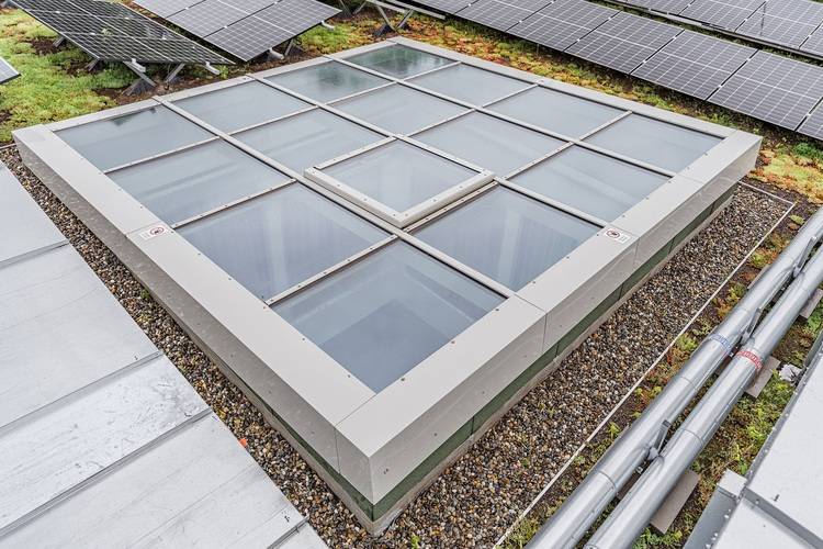 ALT-Text: „Großes Glasdachfenster auf einem Flachdach mit Kieselsteineinfassung, umgeben von modernen Solarpanelen zur nachhaltigen Energiegewinnung in der Gebäudetechnik.