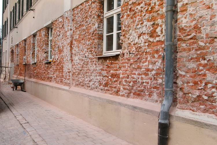 Alt-Text: „Detailansicht einer Gebäudefassade mit freiliegender Ziegelstruktur und sichtbaren Abnutzungsspuren, inklusive mehrerer Fenster. Im Vordergrund befindet sich ein Regenabflussrohr, während im unteren Bereich eine Schubkarre auf einem gepflasterten Weg steht – Symbol für laufende Sanierungsarbeiten und die Relevanz von Instandhaltungsmaßnahmen in der Gebäudeplanung.“.