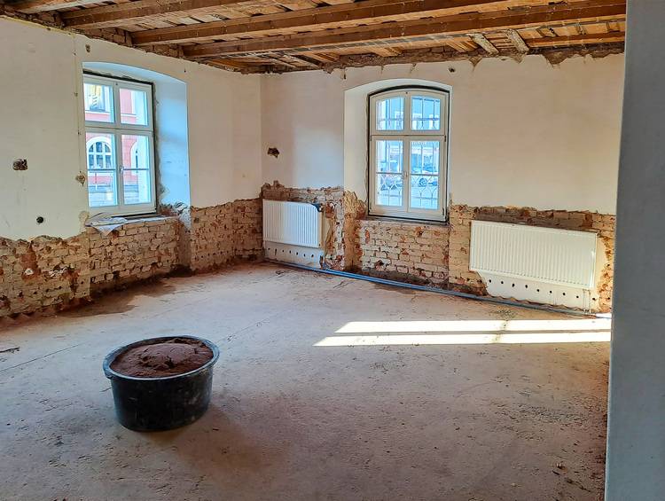 ALT-Text: „Teilweise renovierter Raum mit freiliegenden Ziegelwänden, zwei moderne Fenster für verbesserte Energieeffizienz und ein Eimer als Teil der Bauarbeiten zur Optimierung der Gebäudetechnik.“.