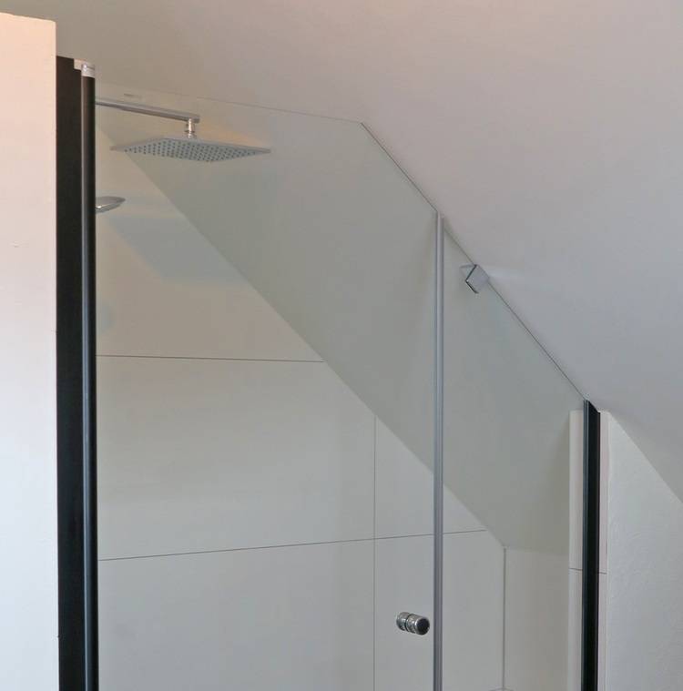 Alt-Text: „Barrierefreies Duschsystem in einem modernen Bad mit geneigter Decke, transparenter Glasabtrennung und großem Regenduschkopf. Weiße Fliesen erhöhen die Lichtreflexion und erleichtern die Reinigung, ideal für energieeffiziente Gebäude- und Sanitärprojekte.“.