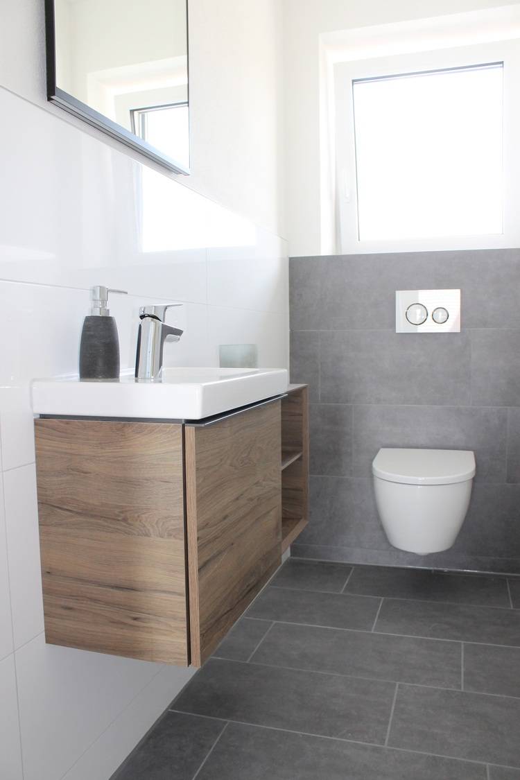 ALT-Text: „Barrierefreies, modernes Badezimmer mit grauen Fliesen und schwimmendem Holzwaschtisch inklusive eingebautem Waschtischarmatur. Wandmontiertes, ergonomisches WC neben einem großen Fenster zur Förderung natürlicher Belichtung.