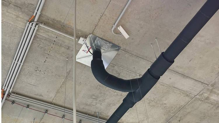 ALT-Text: „Freiliegende Rohre und Leitungen an einer Betondecke mit einem schwarzen, isolierten Lüftungskanal, exemplarisch für moderne Gebäudetechniklösungen in der SHK-Branche.