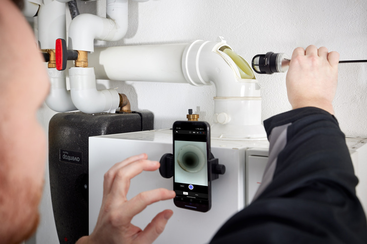 ALT-Text: „Fachhandwerker inspiziert Rohrleitung mit Kamerasonde über Smartphone zur Qualitätskontrolle in der Gebäudetechnik.“.