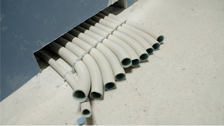 ALT-Text: Graue, flexible Installationsrohre für Elektro- und Datenkabel verlaufen aus einer Wandöffnung über einen hellen Boden, symbolisierend die Integration moderner Gebäudetechnik für effektive Raumplanung.