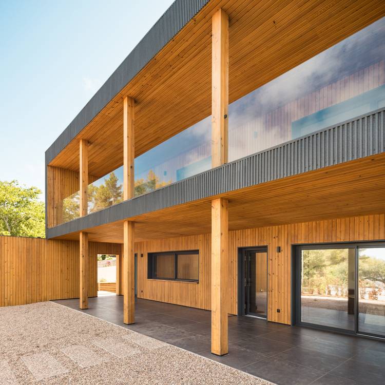 Alt-Text: „Energieeffizientes Holzgebäude mit großer Verglasung und vertikaler Trägerstruktur, umgeben von einem Kiesinnenhof; Beispiel für nachhaltige Architektur in der Gebäudetechnikbranche.