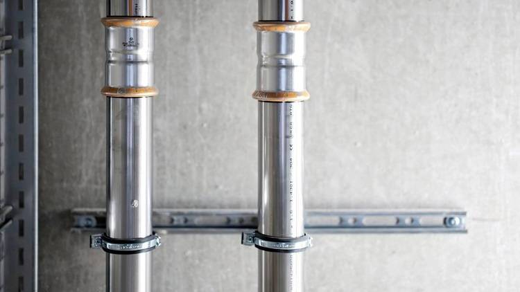 Alt-Text: „Zwei glänzende Edelstahlrohre mit robusten Halterungen montiert an einer grauen Betonwand, repräsentieren moderne Installationslösungen in der Heizungs- und Klimatechnik für Energieeffizienz in Gebäuden.