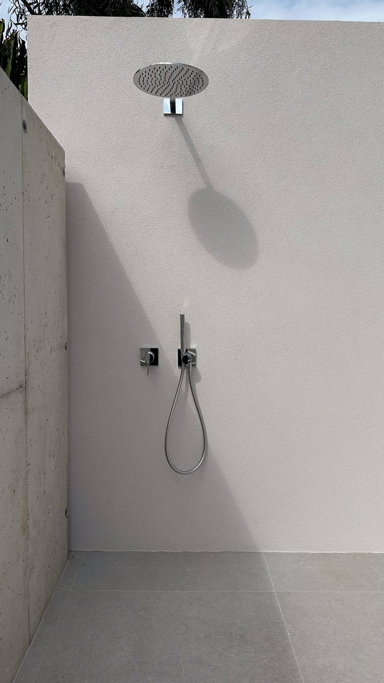 Alt-Text: „Moderne Außendusche mit runder Überkopfbrause und Handbrause, montiert an einer hellen Wand, optimiert für energieeffiziente Wassernutzung im Gebäudedesign.