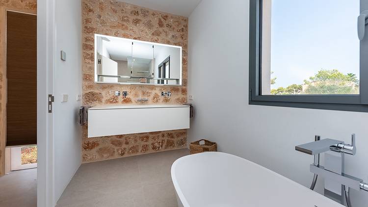 ALT-Text: „Modernes Badezimmer mit freistehender Badewanne, großes Fenster für optimale Tageslichtnutzung, Steinakzentwand für erhöhte Energieeffizienz und Doppelwaschtisch mit Spiegeln – geeignet für barrierefreie Planungen in der gehobenen Gebäudetechnik.