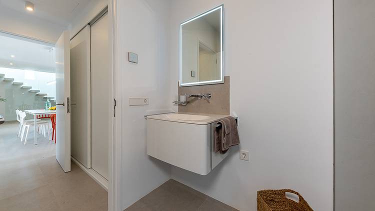 Ein modernes Badezimmer mit wandmontiertem Waschbecken, energiesparendem LED-Spiegel und Handtuchhalter; im Hintergrund ein Einblick in einen offenen Außenbereich für energieeffizientes Wohnen.