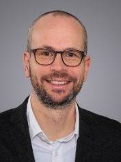 Alt-Text: „Ein lächelnder Gebäudetechnikspezialist in Business-Kleidung vor grauem Hintergrund, symbolisiert Expertise und Professionalität im Bereich Sanitär-, Heizungs- und Klimatechnik.