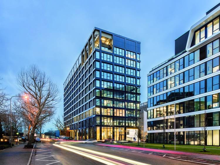 ALT-Text: „Moderne Bürogebäude aus Glas bei Dämmerung mit Lichtspuren vorbeifahrender Fahrzeuge, symbolisierend innovative Gebäudetechnik und energieeffiziente Lösungen im urbanen Umfeld.