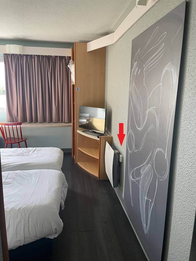 ALT-Text: „Hotelzimmer mit zwei Betten, rotem Stuhl und TV. Ein großes abstraktes Gemälde an der Wand, ergänzt durch eine rote Markierung, die auf einen modernen Luftreiniger hinweist – ideal für Entscheider in der Gebäudetechnik zur Verbesserung von Raumluftqualität und Energieeffizienz.