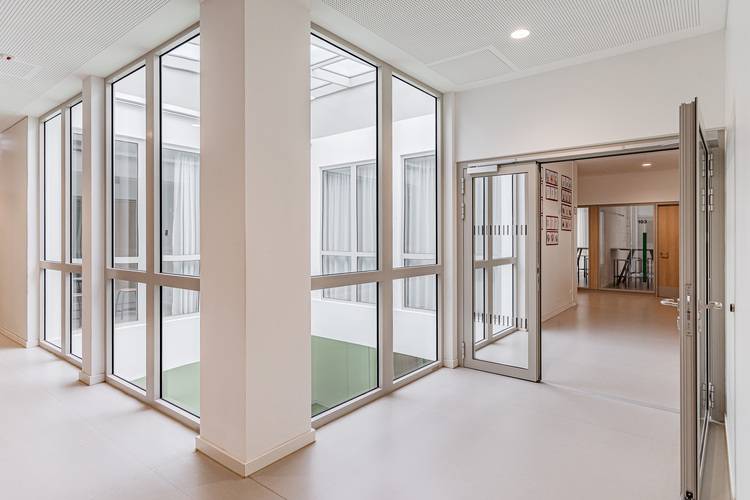 Alt-Text: „Moderner Gebäudeflur mit energieeffizienten, bodentiefen Fenstern und einer offenen Tür zu einem weiteren Korridor, stellvertretend für innovative Klimatechniklösungen in der zeitgenössischen Architektur.“.