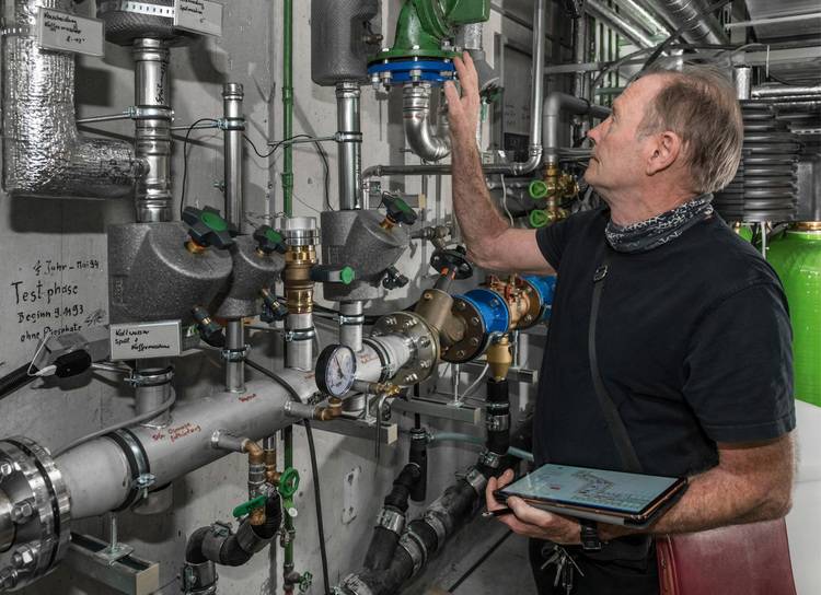 ALT-Text: „Fachmann in einem Technikraum prüft ein Heizsystemventil mithilfe eines Tablets, um die Effizienz der Gebäudetechnik zu optimieren. Umgeben von komplexen Rohrleitungen und Messgeräten zur Unterstützung der Instandhaltungsprozesse.
