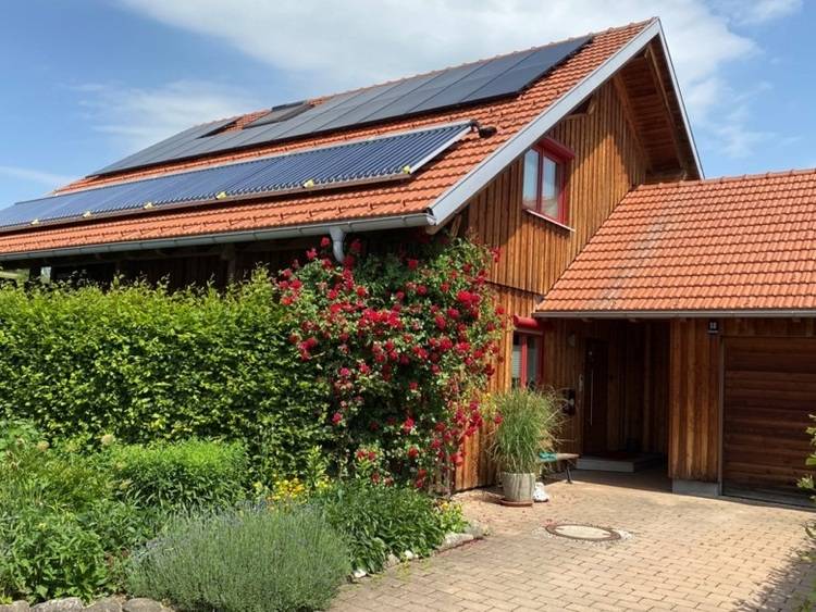 Alt-Text: „Energieeffizientes Gebäude mit Solarpanels auf dem roten Ziegeldach, umgeben von einem nachhaltigen Garten, unterstreicht moderne Lösungen für erneuerbare Energien und innovative Gebäudetechnik.“.