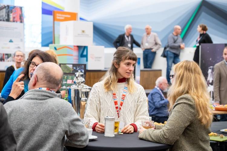 ALT-Text: „Entscheider der Gebäudetechnikbranche im Gespräch an einem runden Tisch während einer Fachmesse; im Hintergrund Informationsstände zu neuesten Trends in der Sanitär-, Heizungs- und Klimatechnik.
