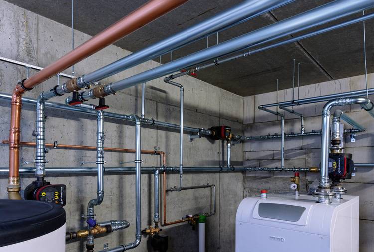 Alt-Text: „Detailaufnahme einer modernen Heizungs- und Sanitärinstallation in einem Betonuntergeschoss, einschließlich vernetzter Rohre und eines hocheffizienten Gas-Brennwertkessels, als Beispiel für fortschrittliche Gebäudeenergiesysteme.