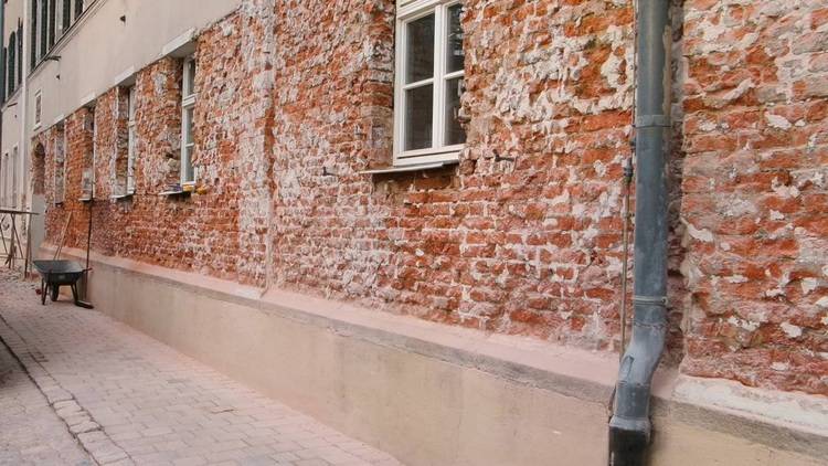 Alt-Text: „Eine historische Backsteinmauer mit abblätterndem Putz, kleinen Fenstern und einem Regenabflussrohr entlang eines schmalen Kopfsteinpflasterwegs, repräsentiert Herausforderungen und Potenziale bei der energetischen Sanierung von Gebäudehüllen in der Gebäudetechnikbranche.“.