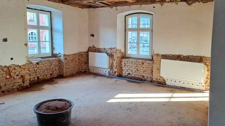 Alt-Text: „Raum im Rohbauzustand mit freigelegten Ziegelwänden, zwei Fenstern zur verbesserten Wärmedämmung und einem Farbeimer auf dem Boden, symbolisiert den Fortschritt in der energieeffizienten Gebäuderenovierung.“.