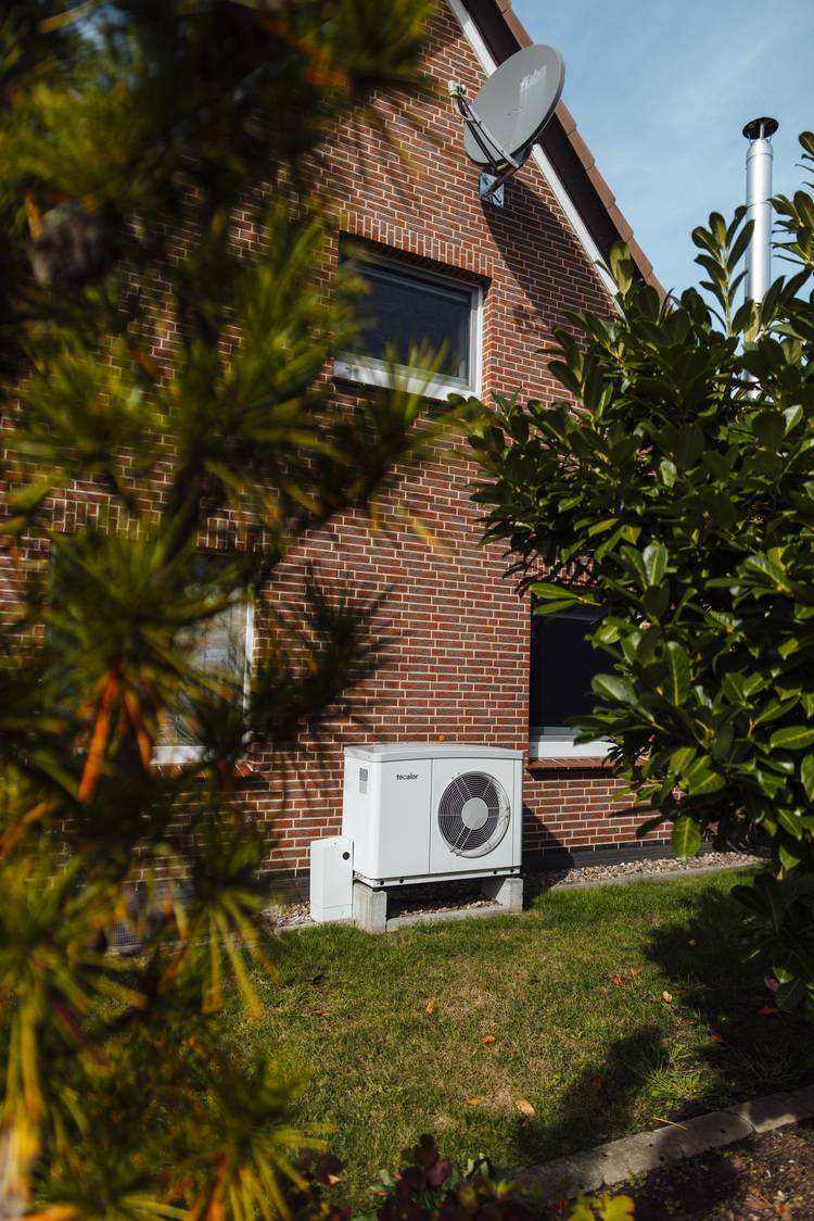 Alt-Text: „Außenklimagerät an der Ziegelwand eines Wohnhauses, umgeben von Pflanzen; ideale Lösung für energieeffiziente Kühlung im Gebäudetechniksektor.