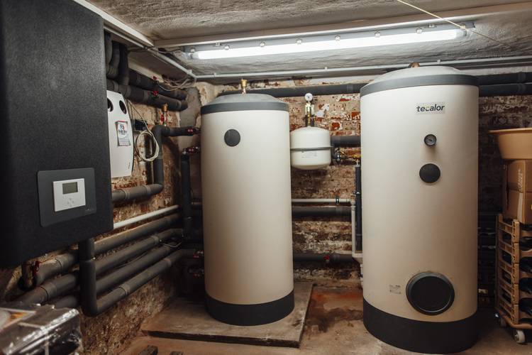 Sicht auf zwei große, zylindrische Wasserspeicher und Rohrleitungen in einem Keller mit freiliegenden Ziegelwänden, ausgestattet mit einem Steuerschrank – effiziente Heizlösungen für moderne Gebäudetechnik.