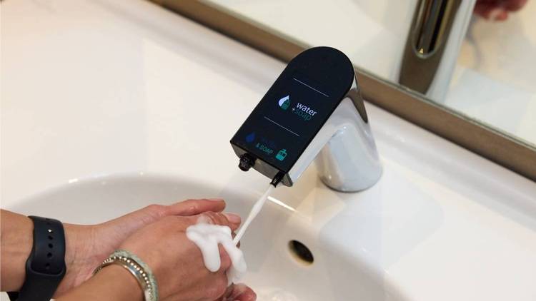 Alt-Text: „Hände, die unter einem modernen Wasserhahn mit digitalem Wasserspargerät und Echtzeitanzeige der Wasserverbrauchsdaten gewaschen werden; symbolisiert innovative Sanitärtechnik für Energieeffizienz in Gebäuden.“.