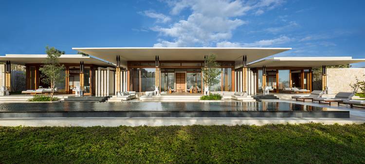 ALT-Text: "Moderne Villa mit großen, energieeffizienten Fenstern und nachhaltiger Gebäudetechnik, umgeben von Begrünungskonzepten für optimierte Energieeffizienz. Reflektierendes Wasserbecken im Vordergrund als Teil des Wärmemanagements. Blauer Himmel zeigt optimalen Einsatz erneuerbarer Energien.