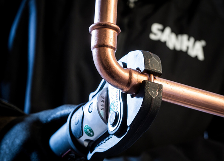 ALT-Text: „Ein Fachhandwerker nutzt ein Presswerkzeug, um eine Kupferrohrverbindung in einem SHK-Projekt zu crimpen. Der Handwerker trägt eine schwarze SANHA-Jacke und befindet sich auf einer Baustelle für Heizungs- und Klimatechnik.