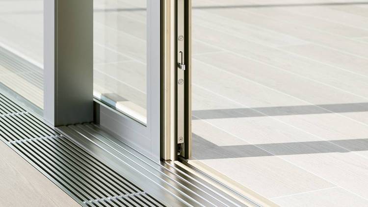 ALT-Text: „Teilgeöffnete Schiebetür aus Glas mit metallener Bodenschiene und hellem Holzfußboden, ideal für moderne Gebäude mit Fokus auf Energieeffizienz und Barrierefreiheit in der Gebäudetechnik.