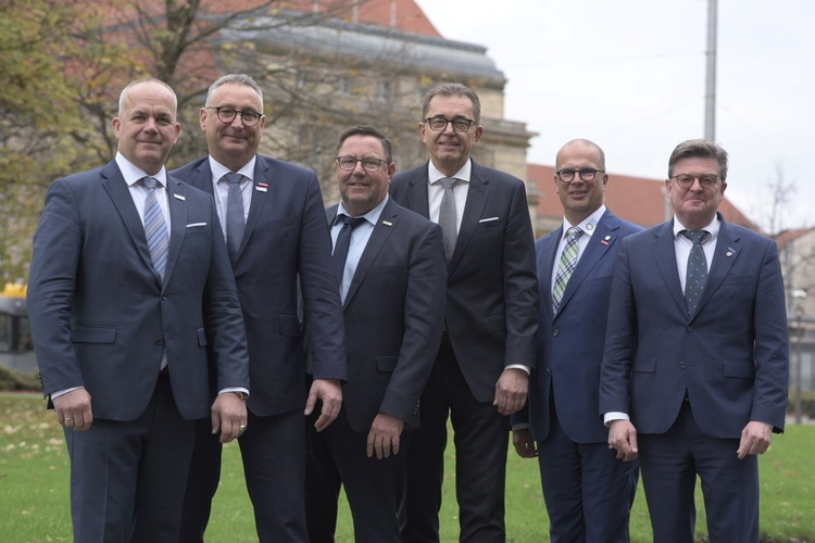 Alt-Text: „Sechs Geschäftsleute in Anzügen auf einer grünen Rasenfläche vor einem modernen Gebäude diskutieren über nachhaltige Gebäudetechniklösungen.