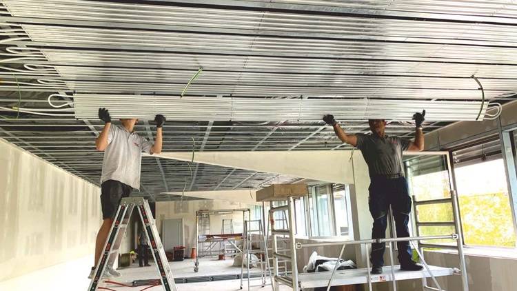 Alt-Text: „Zwei Fachhandwerker installieren akustisch optimierte Metallpaneele an der Decke eines neuen Bürogebäudes. Sie nutzen Leitern und Gerüste für den Zugang, um die energieeffiziente Raumlufttechnik zu integrieren.“.