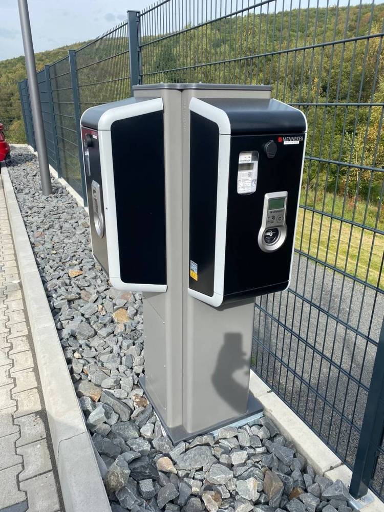 Alt-Text: „Zwei schwarze Ladestationen für Elektrofahrzeuge auf gepflasterter Fläche, mit Metallzaun und Grünanlagen im Hintergrund; Fokus auf energieeffiziente Infrastruktur in der Gebäudetechnik.