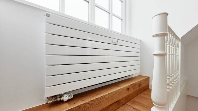 Alt-Text: „Moderner weißer Wandheizkörper in energetisch effizientem Design, montiert auf stilvollen Holzstufen neben einem Treppengeländer aus Metall und großen Fenstern, die Tageslicht hereinlassen – Beispiel für innovative Heiztechnik im Gebäudedesign für TGA-Planer und Architekten.“.