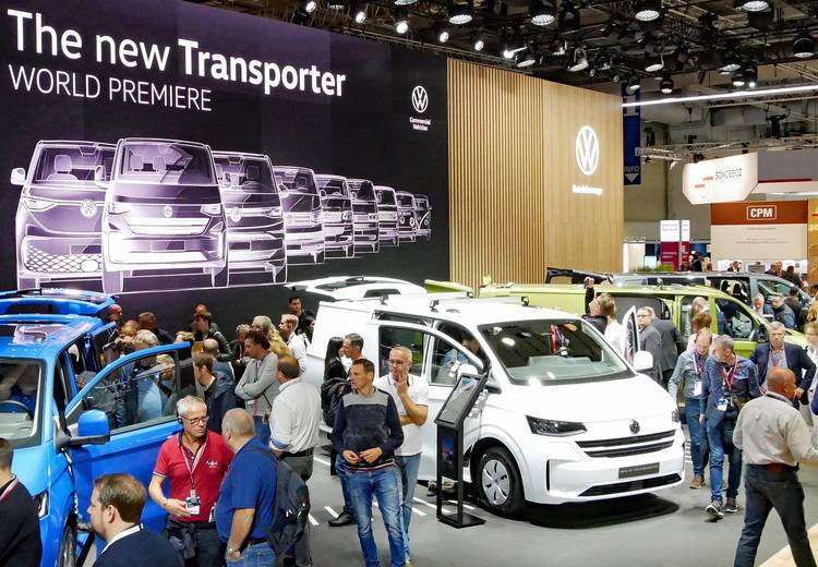 Alt-Text: „Fachbesucher auf einer Gebäudetechnikmesse betrachten innovative Transportermodelle von Volkswagen, präsentiert vor einem thematischen Hintergrund für erneuerbare Energien und nachhaltige Mobilitätslösungen.