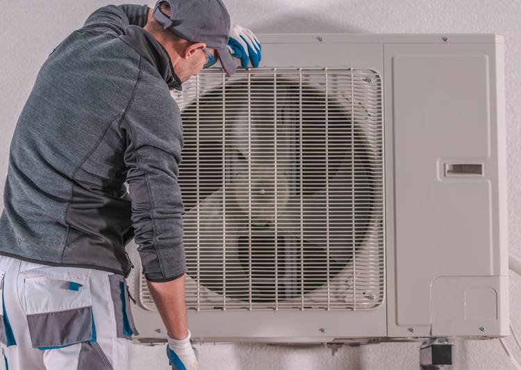 Alt-Text: „Fachhandwerker in Arbeitskleidung bei der Wartung einer wandmontierten Außeneinheit für Heizungs-, Lüftungs- und Klimatechnik (HVAC) im Rahmen energieeffizienter Gebäudetechnik.