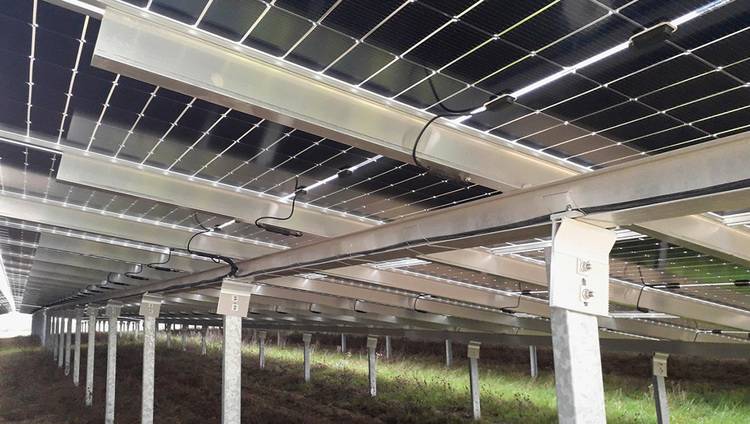 ALT-Text: "Untersicht einer Solaranlagenanordnung mit Metallrahmen auf grasbewachsenem Boden, detailliert dargestellte Verkabelung und Trägerstrukturen, veranschaulicht die Installationstechniken zur Optimierung der Energieeffizienz in der Gebäudetechnik.