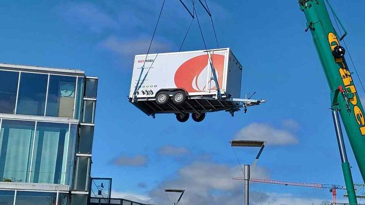 Alt-Text: „Ein Kran hebt einen weißen Sanitärcontainer mit rotem Firmenlogo, der bei der Installation modernster Klimatisierungstechnik auf einer Baustelle zum Einsatz kommt, unter blauem Himmel.“.
