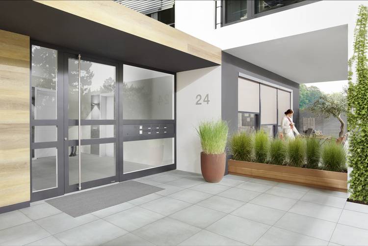 Alt-Text: „Moderner Gebäudeeingang mit automatischen Glastüren und energieeffizienter Fassade, ergänzt durch barrierefreie Zugänge und nachhaltige Begrünung; im Vordergrund ein Fachhandwerker der Gebäudetechnik bei der Wartung.“.