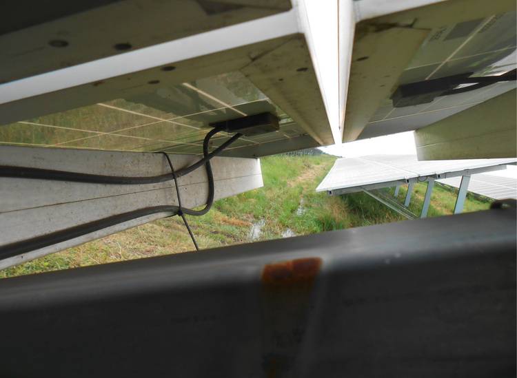 Alt-Text: „Blick unter Solarmodule mit sichtbaren Verkabelungen und Metallstützen, installiert über einer Rasenfläche als Teil eines energieeffizienten Gebäudetechnikprojekts.“.