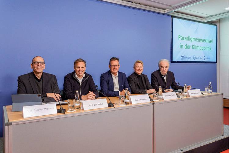 Alt-Text: „Fünf Fachleute aus der Gebäudetechnikbranche diskutieren an einem Konferenztisch, während auf einem Bildschirm die Präsentation ‚Paradigmenwechsel in der Klimapolitik‘ gezeigt wird. Die Teilnehmer sind Entscheidungsträger und Experten für erneuerbare Energien und Energieeffizienz.“.
