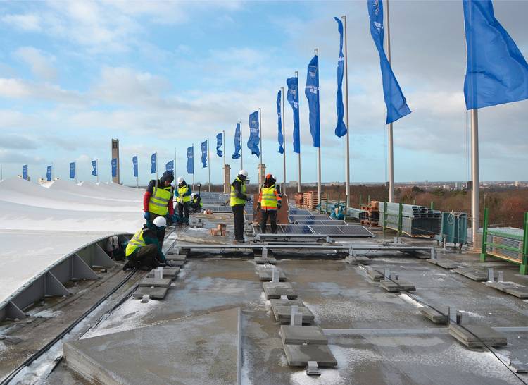 Alt-Text: „Fachkräfte in Sicherheitswesten begutachten eine mit Solarmodulen bedeckte Dachfläche, umgeben von blauen Markierungen, zur Optimierung der Energieeffizienz in der Gebäudetechnik.“.